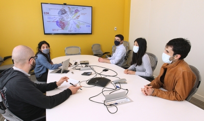 Yuri Pritykin and his lab group sitting around table in a conference room. They discuss a slide with a cluster graph on the TV monitor on the wall.