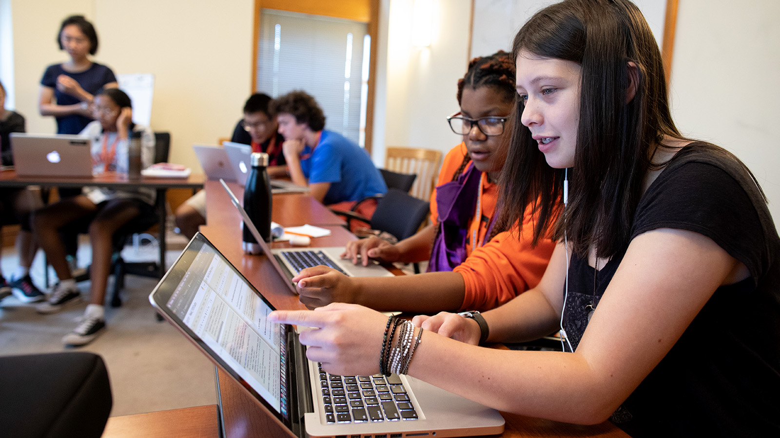 Two high school age girls pointing at their laptop screens in a classroom of students.