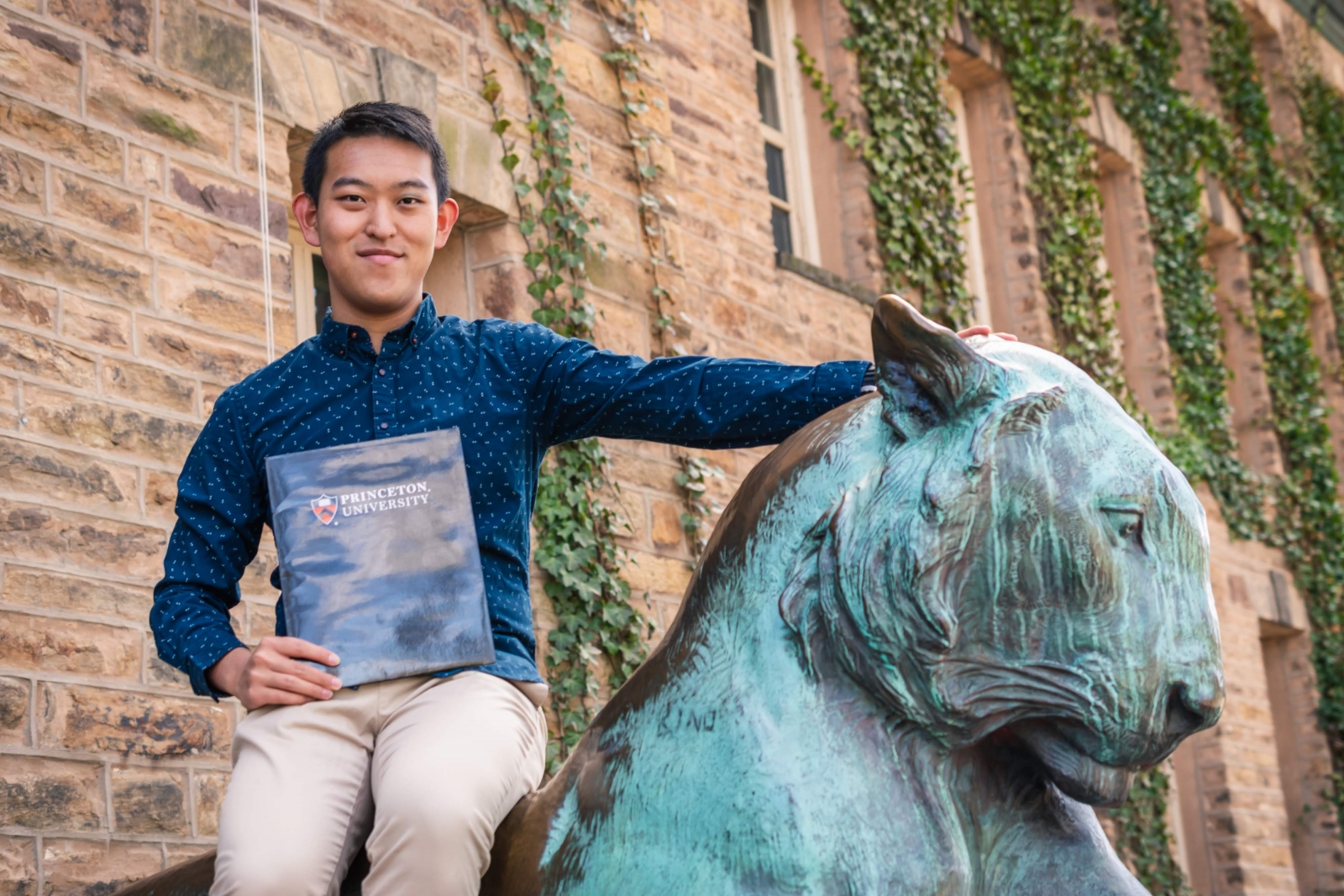 Yang Song sitting on the bronze tiger on the front steps of Nassau Hall. He's holding a black folder with the Princeton University logo on the front.